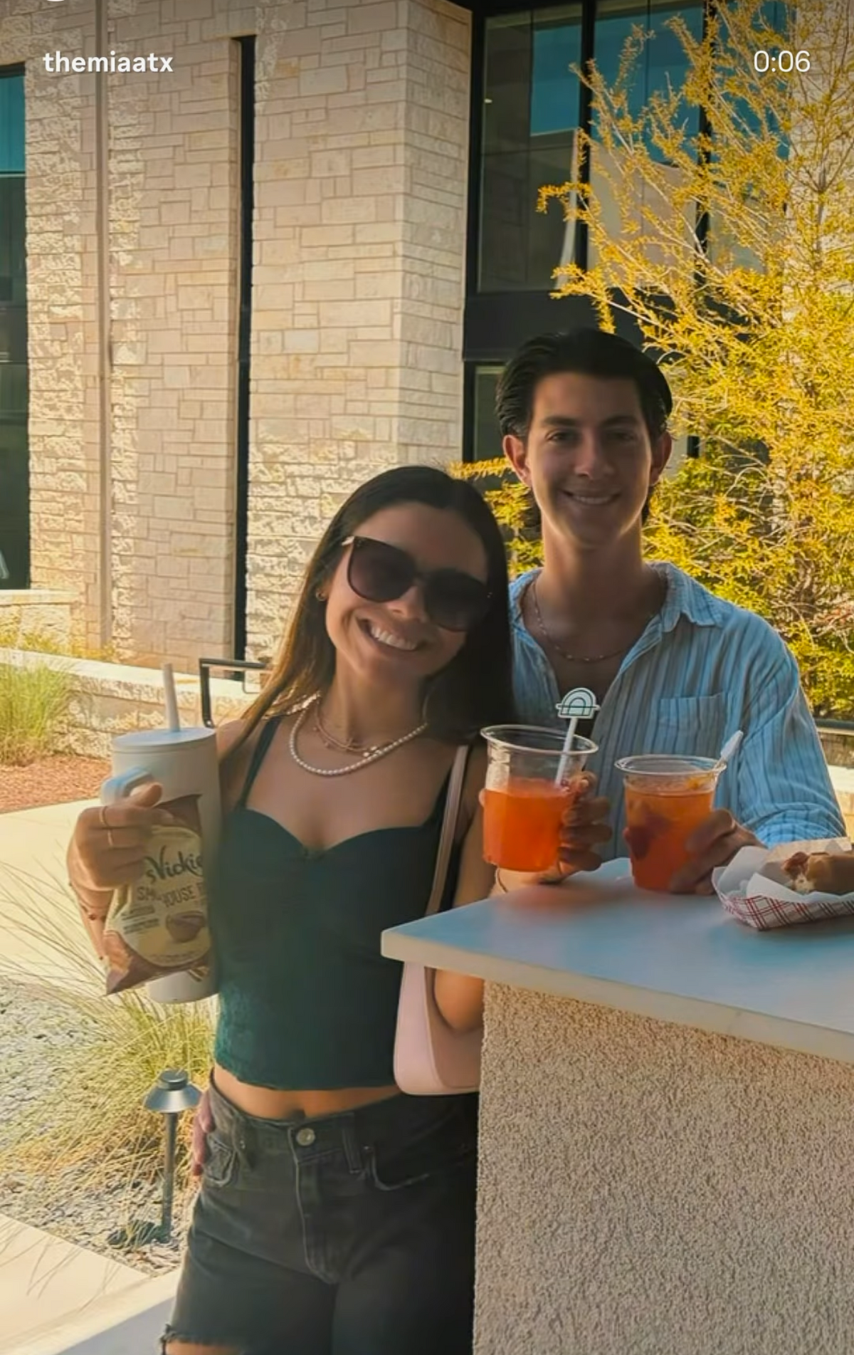 A smiling woman in a dark green crop top and black shorts stands next to a man in a light blue button-down shirt, holding drinks and a bag of Nectar energy drinks outdoors in front of a modern building with stone walls.