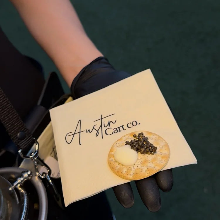 A hand wearing a black glove holds a square cream-colored card or napkin printed with the script logo 'Austin Cart co.' and a small food photo.
