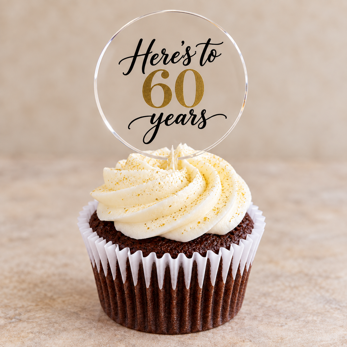 Clear round acrylic cupcake topper reading “Here's to 60 years” in black and gold on a chocolate cupcake with white icing and gold edible glitter.