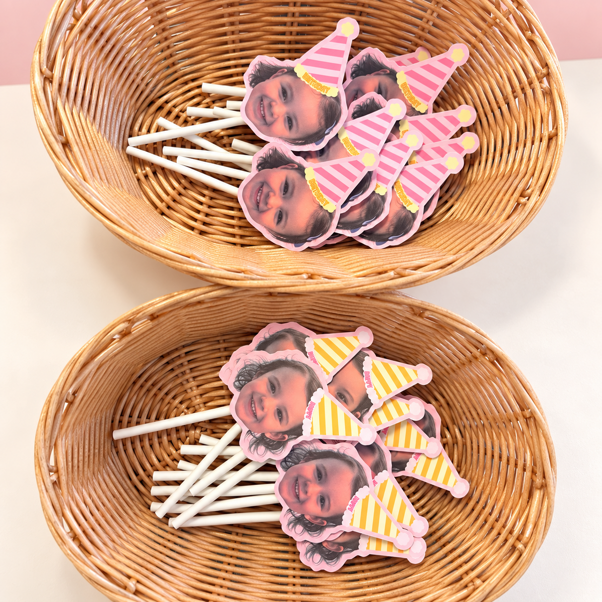 Personalized photo cupcake toppers with pink and yellow striped birthday hats arranged in two wicker baskets.