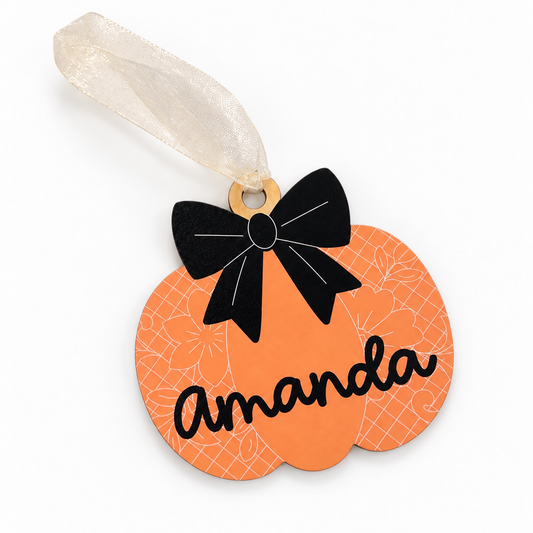 Angled view of orange floral pumpkin tag with black bow, Amanda name and cream ribbon.
