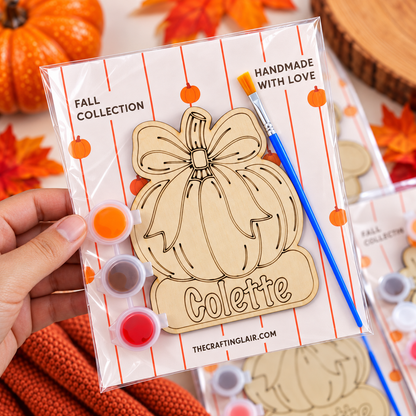 A hand holds a packaged wooden pumpkin-shaped craft piece labeled 'Colette' with paint pots and a paintbrush in a fall-themed setting. The package text includes 'FALL COLLECTION' and 'HANDMADE WITH LOVE,' and the brand 'THECRAFTINGLAIR.