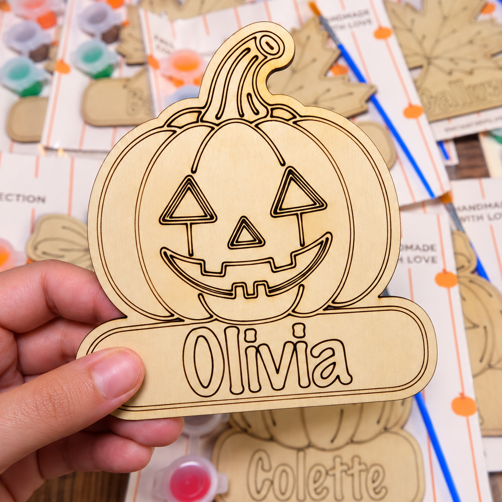 A hand holds a laser-cut natural wood pumpkin sign engraved with a jack-o'-lantern face and the name 'Olivia'. The background shows a crafting table covered with similar wooden sign templates and paint palettes.