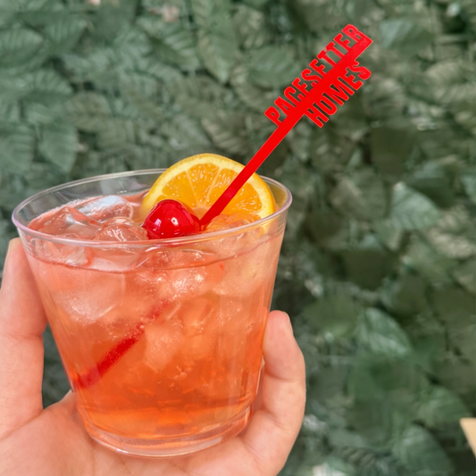 A hand holding a clear plastic cup filled with an orange-pink iced cocktail, garnished with a yellow orange slice and a red cherry, with a red plastic straw that reads 'PASCUALINO HOMMES' against a blurred green leaf wall background.