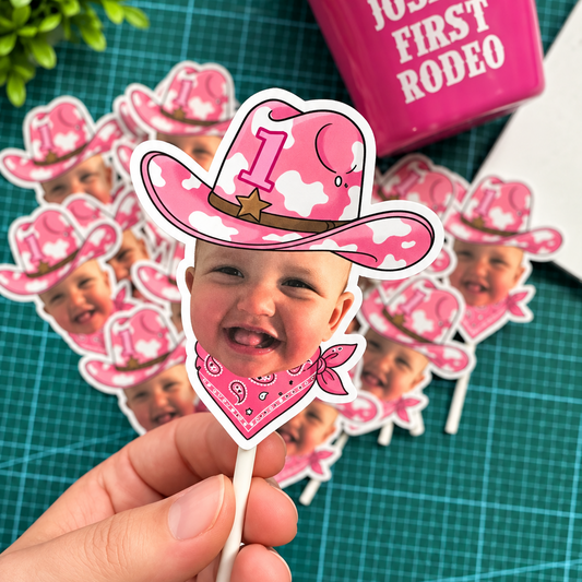 A hand holds a die-cut cowgirl-themed first birthday topper with a baby photo insert, pink camouflage hat, and pink bandana. More matching toppers are scattered on a green cutting mat, with a pink cup in the background.