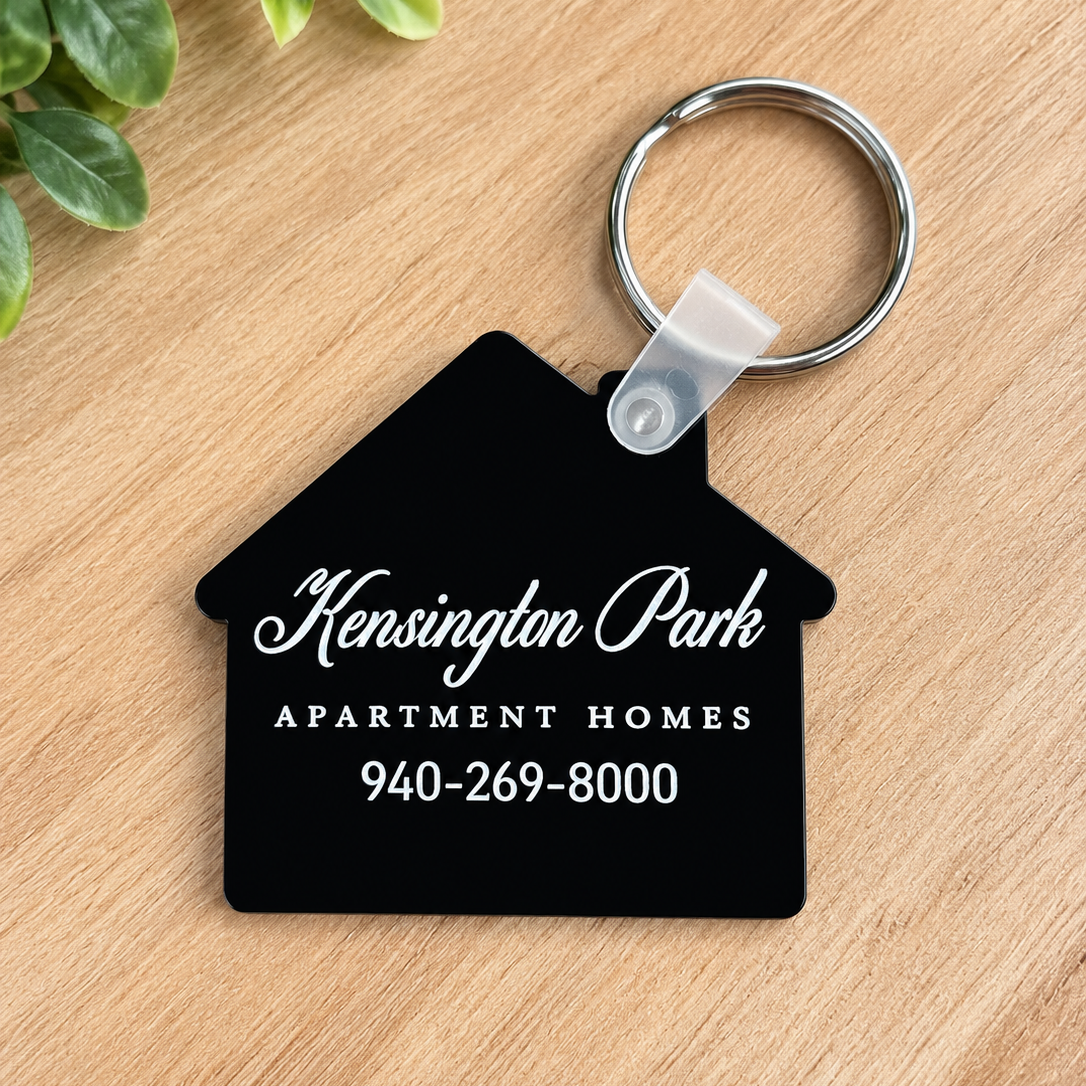 A black house-shaped keychain with white text reading 'Kensington Park Apartment Homes 940-269-8000' attached to a silver key ring, placed on a light wood surface with green leaves in the corner.