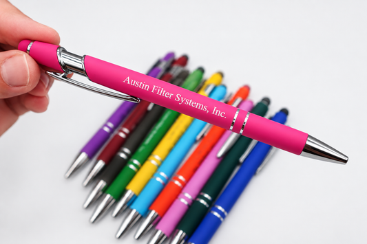 A hand holds a pink retractable pen with silver metal accents and white printed text reading 'Austin Filter Systems, Inc.' in the foreground, with several other colorful pens arranged in a blurred row behind on a white background.