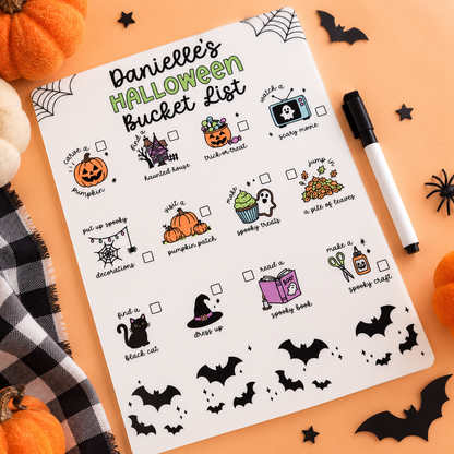 A Halloween-themed planner page titled 'HALLOWEEN Bucket List' laid flat on an orange surface, surrounded by pumpkins, black spider decorations, a black marker, and a black-and-white plaid cloth. The page includes illustrated checklist items such as carving a pumpkin, haunted house trivia, jump scare, spooky drinks, a pile of leaves, put up spooky decorations, a pumpkin patch, spooky treats, a ghost cupcake, a pile of leaves, find a black cat, dress up, read a spooky book, and make a spooky craft.