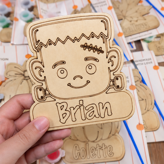 A hand holds a personalized laser-cut wooden Frankenstein sticker template engraved with the name 'Brian'. The background shows a table covered with other wooden templates, paint palettes, and craft supplies.