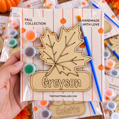 A hand holds a packaged wooden name plaque kit with an engraved maple leaf and the name 'Greyson.' The package includes small paint pots, a blue paintbrush, and autumn-themed packaging.