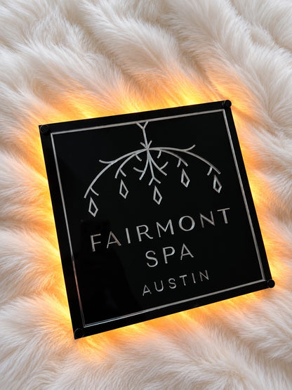 Backlit black acrylic Fairmont Spa logo sign resting on white faux fur.