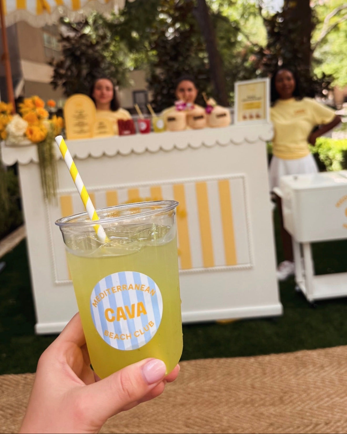 A hand holds a clear plastic cup filled with a yellow beverage and a yellow-and-white striped straw in an outdoor beach club setting. The cup has a circular blue-and-white label reading 'MEDITERRANEAN CAVA BEACH CLUB.