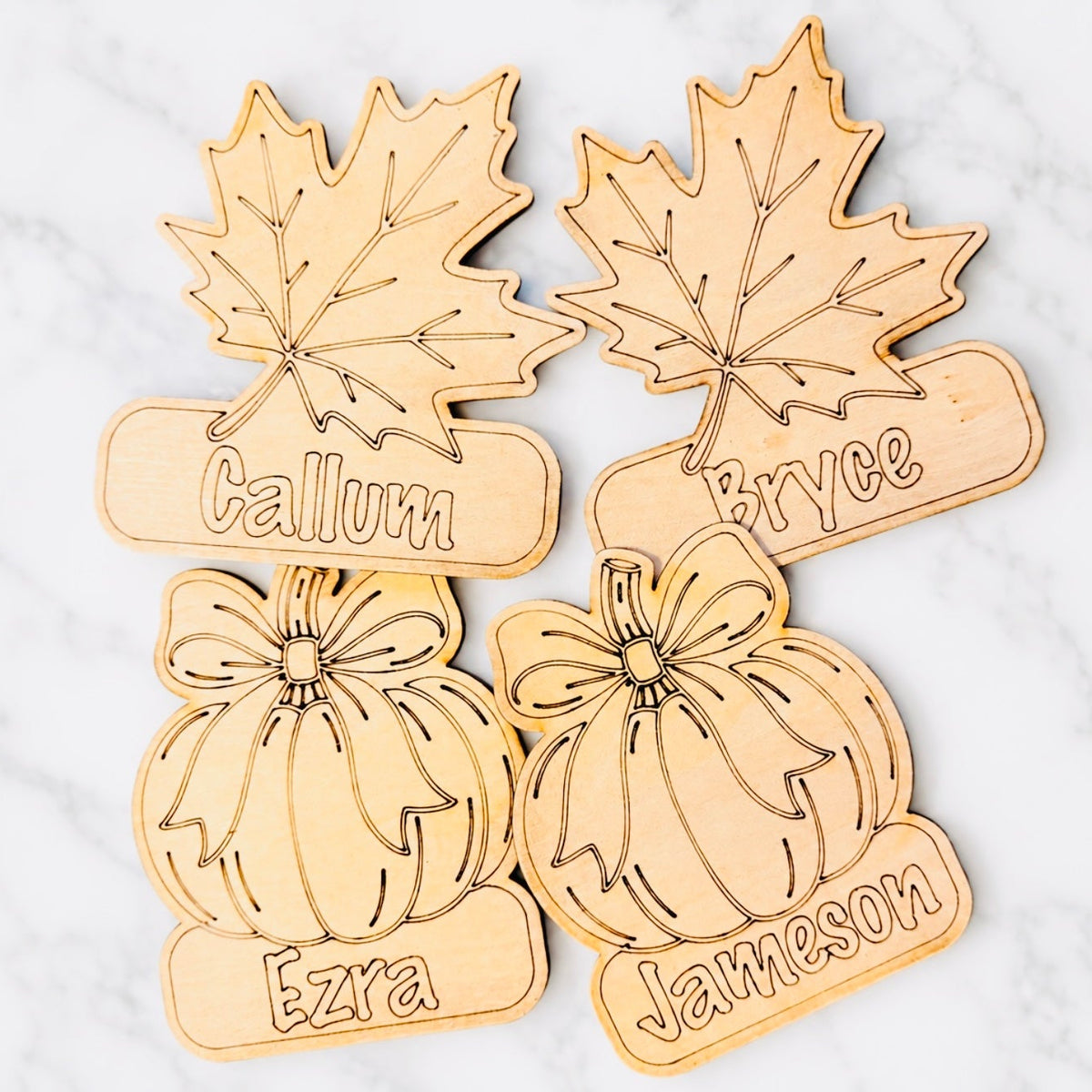Four laser-cut wooden fall-themed name signs featuring maple leaves and pumpkins, each engraved with a personalized name, arranged on a white marble background.