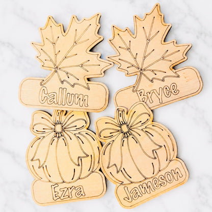 Four laser-cut wooden fall-themed name signs featuring maple leaves and pumpkins, each engraved with a personalized name, arranged on a white marble background.