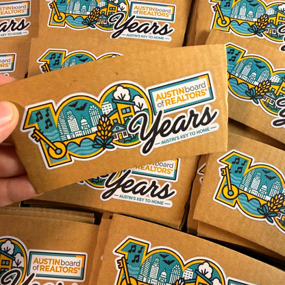 A hand holds a brown kraft paper box printed with a colorful '100 Years Austin Board of Realtors' logo, surrounded by multiple identical boxes on a dark surface.