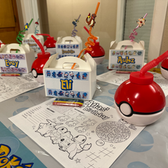 A table set up for a Pokémon-themed birthday party, featuring red and white Pokémon ball-shaped drink cups with red straws, custom white favor boxes with blue and yellow character designs, coloring sheets, and crayons.