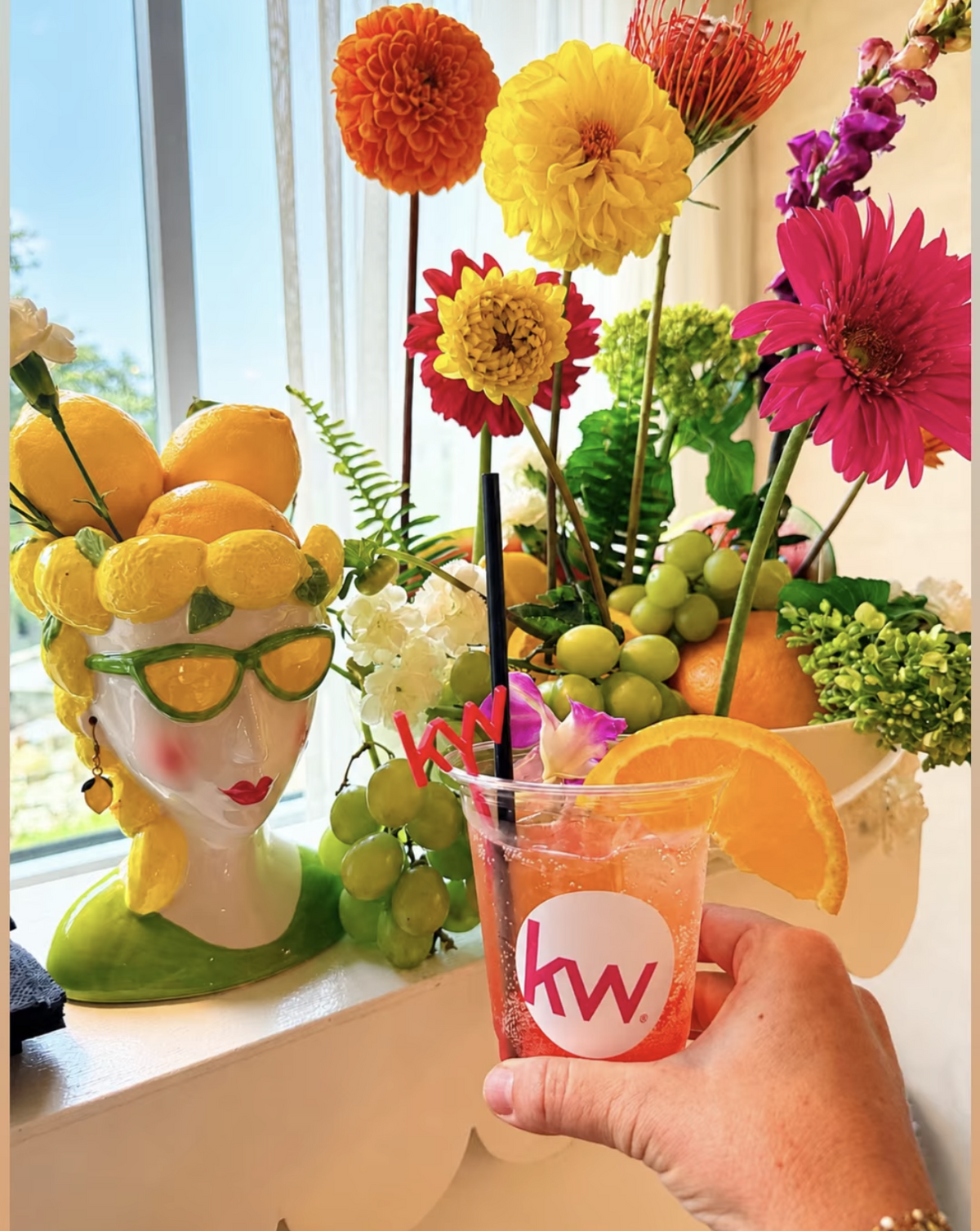 A hand holds a pink drink in a clear plastic cup with a black straw and a white circular logo reading 'hw', set against a bright floral arrangement and a decorative yellow-gloved bust near a window.