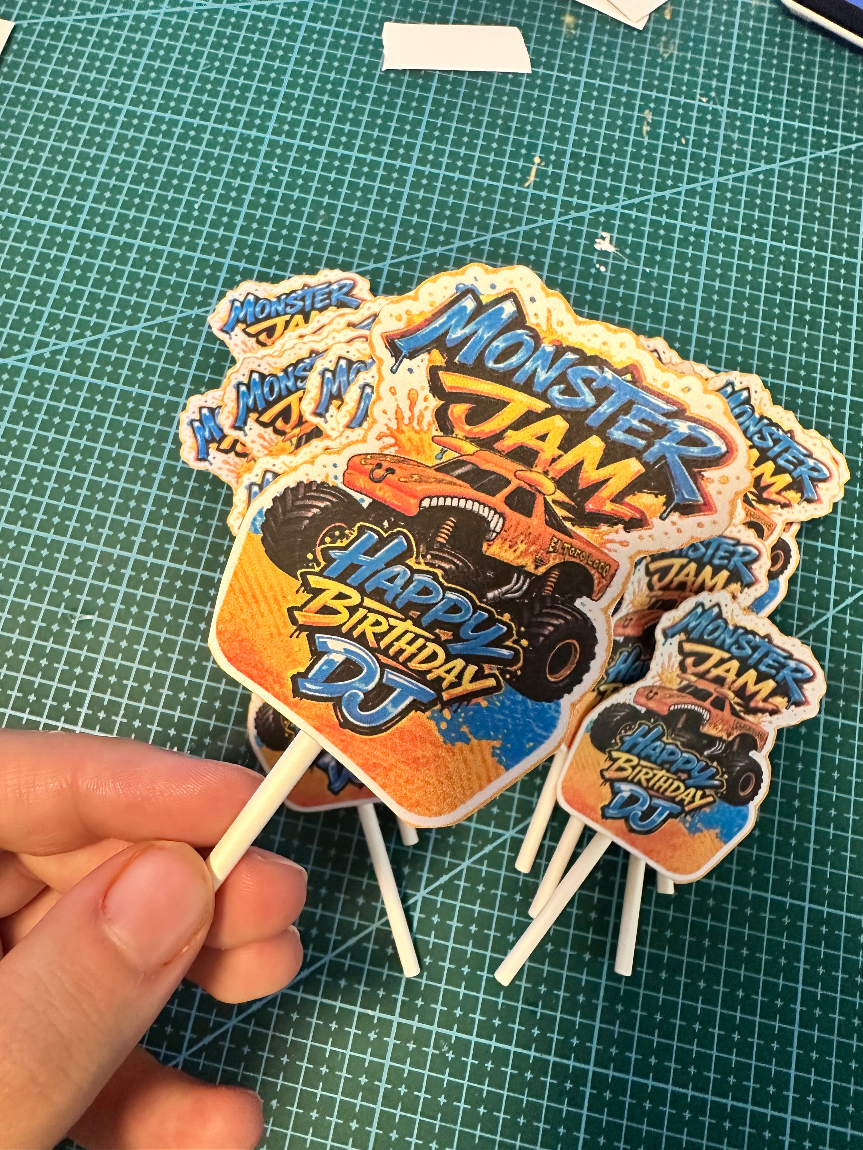 A hand holding a cluster of custom die-cut monster truck birthday cake toppers on wooden sticks, featuring 'Monster Jam' and 'Happy Birthday DJ' designs, set against a green cutting mat background.
