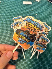 A hand holding a cluster of custom die-cut monster truck birthday cake toppers on wooden sticks, featuring 'Monster Jam' and 'Happy Birthday DJ' designs, set against a green cutting mat background.