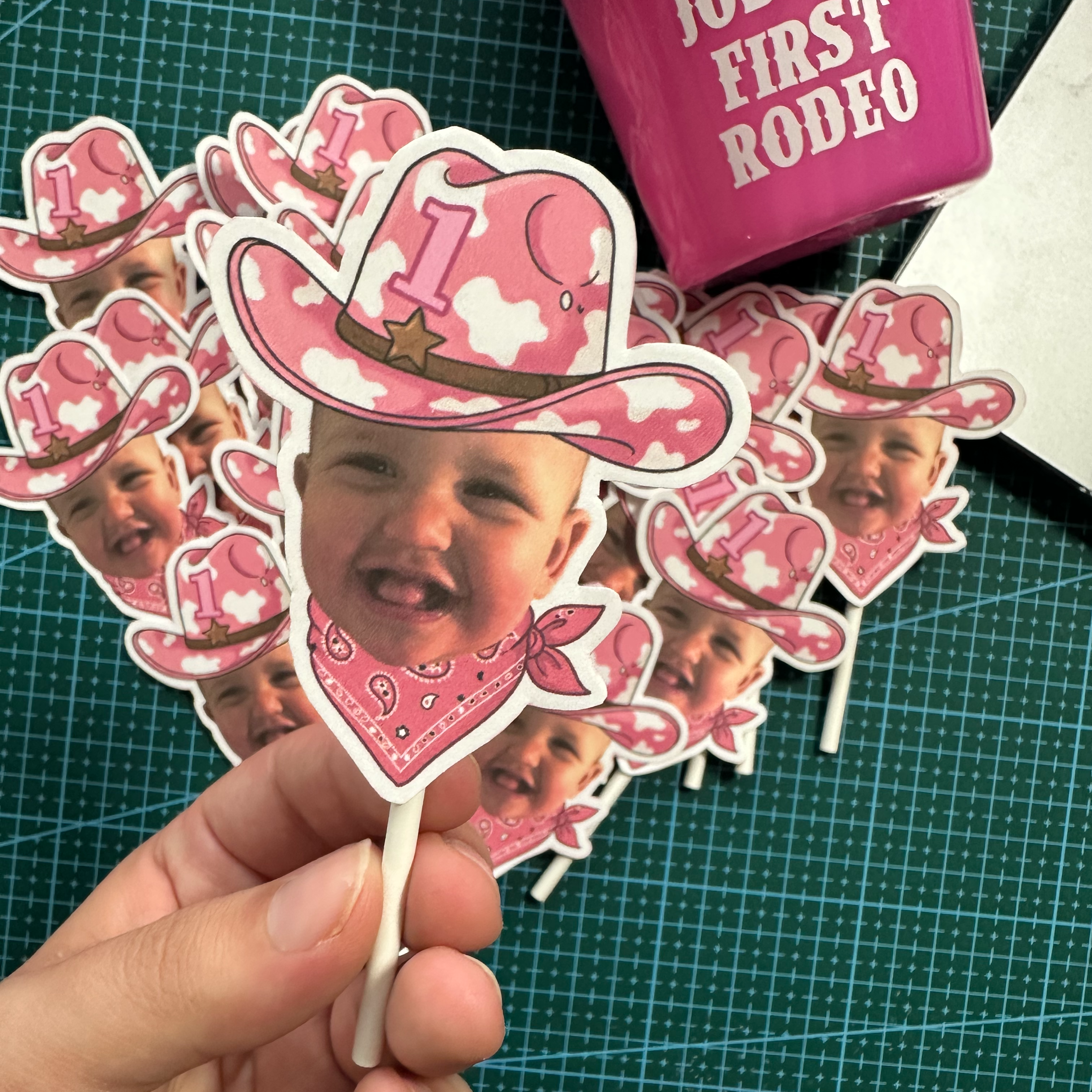 A hand holds a die-cut sticker featuring a baby's face in a pink cowboy hat and bandana, with a pile of similar pink cowboy-themed stickers on a green cutting mat. A pink box labeled 'JOSHUA FIRST RODEO' is partially visible on the left.