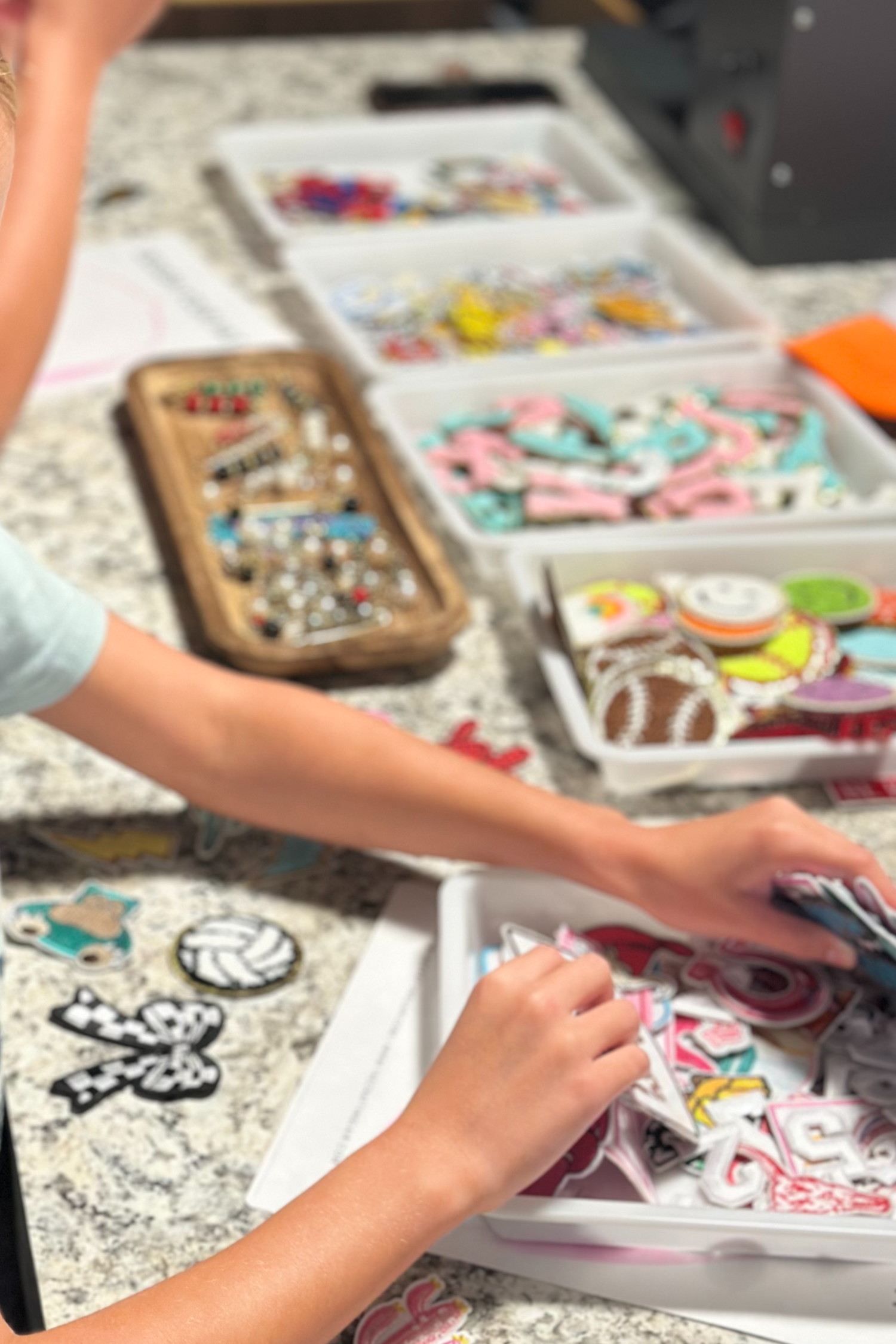 A young girl with blonde hair is shown from the side, carefully sorting through colorful embroidered patches in a clear plastic bin on a granite countertop. Several other bins filled with various embroidered patches are arranged on the counter next to her.