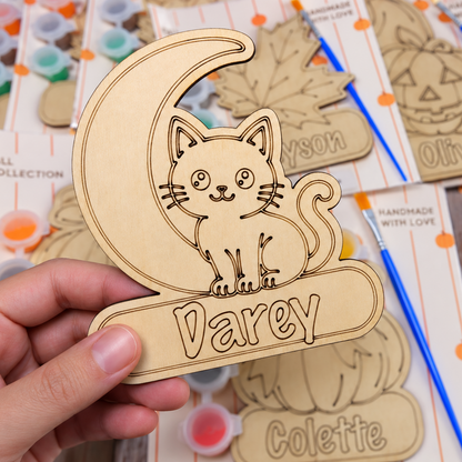 A hand holds a laser-cut wooden name plaque shaped like a crescent moon and cat, engraved with the name 'Darey'. The background shows multiple similar wooden name art pieces in retail packaging, including jack-o'-lantern and autumn-themed designs, along with paintbrushes and small paint pots.