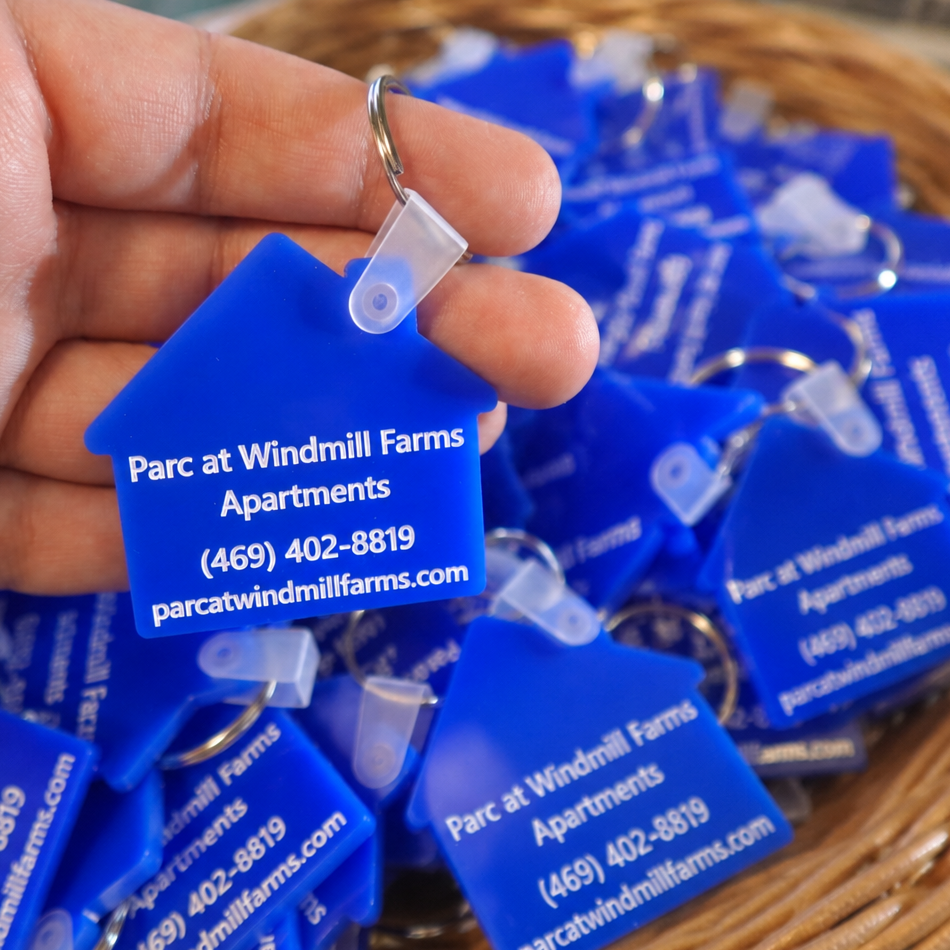 A hand holds a bright blue house-shaped keychain tag printed with the address and contact information for Parc at Windmill Farms Apartments. Multiple matching blue keychain tags are piled in a wicker basket in the background.