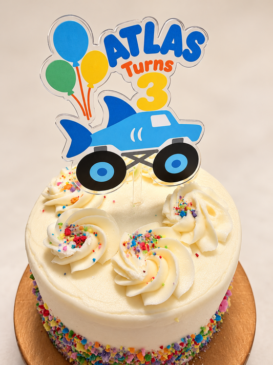 Personalized Atlas Turns 3 blue shark monster truck acrylic cake topper on a birthday cake, elevated close-up