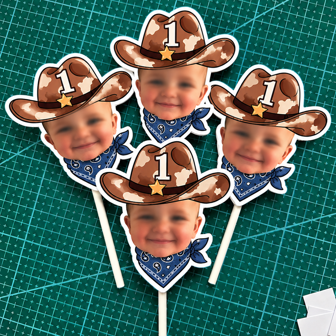 Five cowboy-themed photo popper stands on a green cutting mat. Each stand features a baby's face wearing a brown cowboy hat with a white star, a blue bandana, and a white number 1 on the hat.