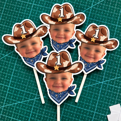 Five cowboy-themed photo popper stands on a green cutting mat. Each stand features a baby's face wearing a brown cowboy hat with a white star, a blue bandana, and a white number 1 on the hat.