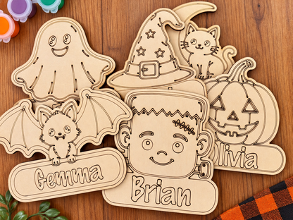 A set of personalized wooden laser-cut Halloween decorations, including a bat, ghost, witch hat, jack-o'-lantern, cat, and names like Emma, Brian, and Olivia, arranged on a wooden tabletop.