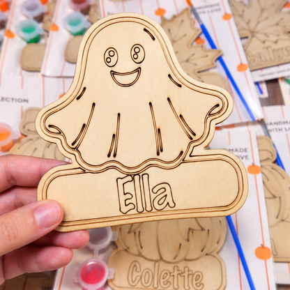 A hand holds a laser-cut wooden ghost ornament labeled 'Ella' in front of a background of other wooden ornaments and craft supplies.