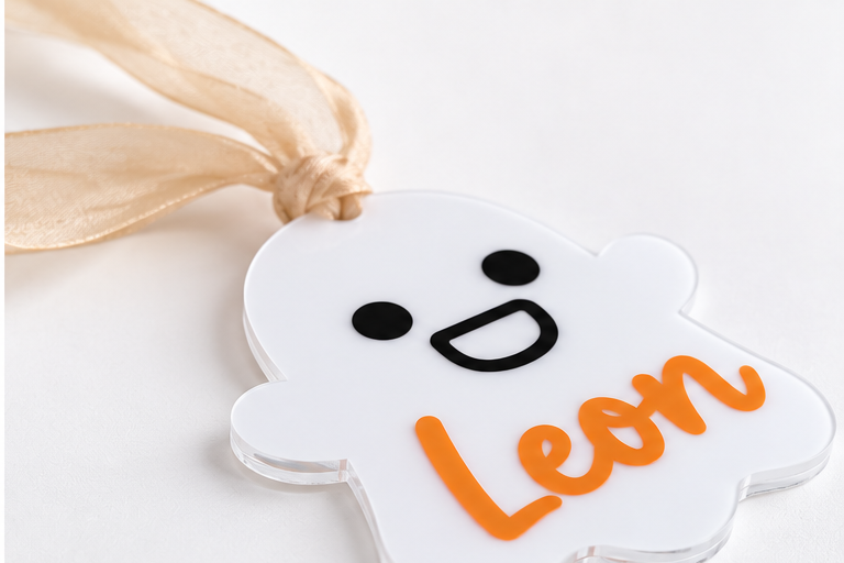 Angled view of personalized white acrylic Halloween ghost name tag with orange Leon lettering and beige ribbon.