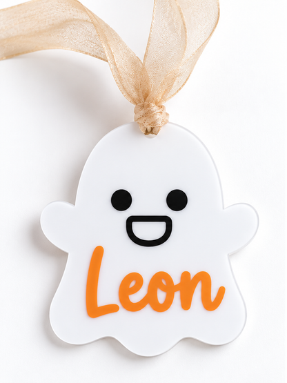 Close-up of white ghost-shaped Halloween name tag with black smiling face, orange Leon lettering and beige ribbon.