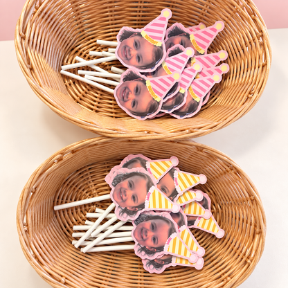 Personalized photo cupcake toppers with pink and yellow striped birthday hats arranged in two wicker baskets.
