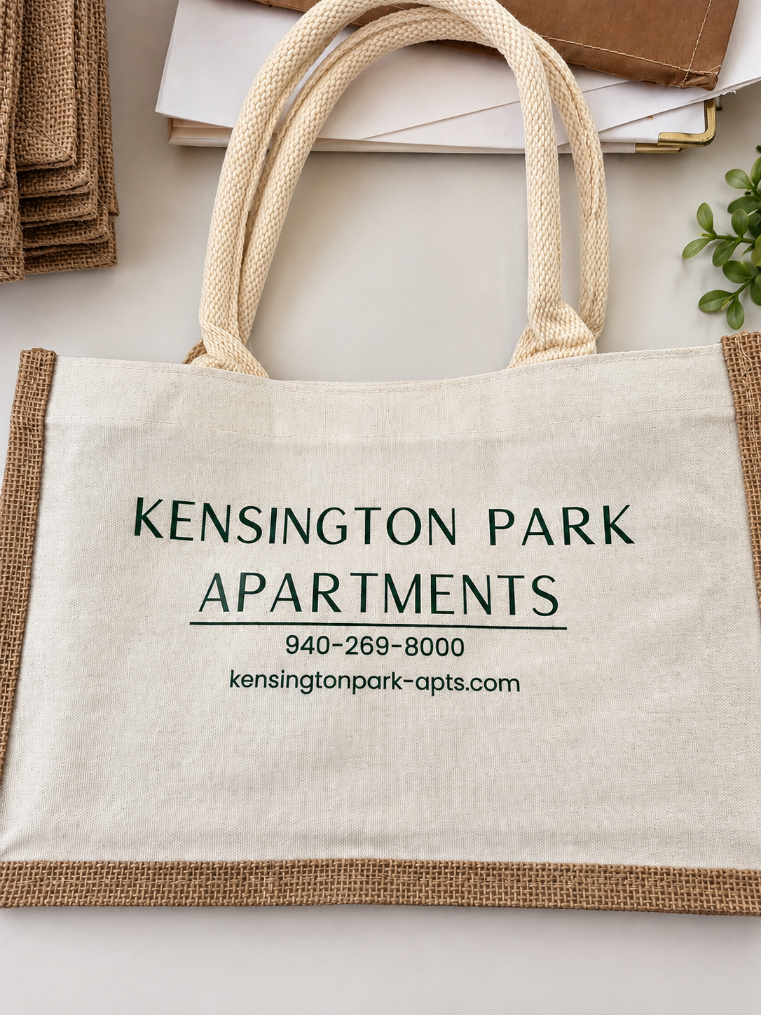 A canvas tote bag with natural jute trim and handles, printed with 'KENSINGTON PARK APARTMENTS' and contact information in dark green text, shown on a light neutral background with styled props.