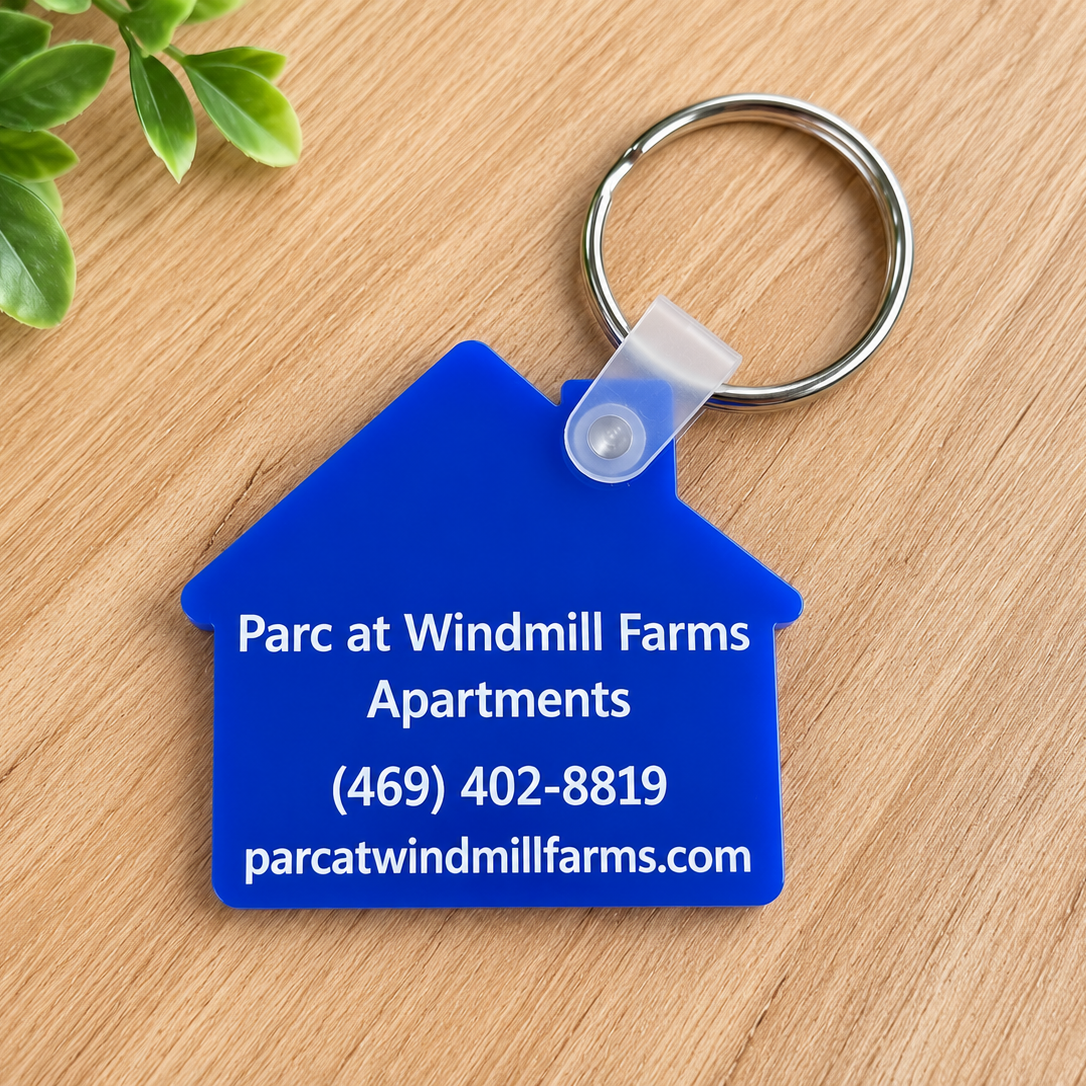 A blue house-shaped acrylic keychain with white text reading 'Parc at Windmill Farms Apartments' and contact details, attached to a silver key ring on a light wood surface with green leaves in the corner.