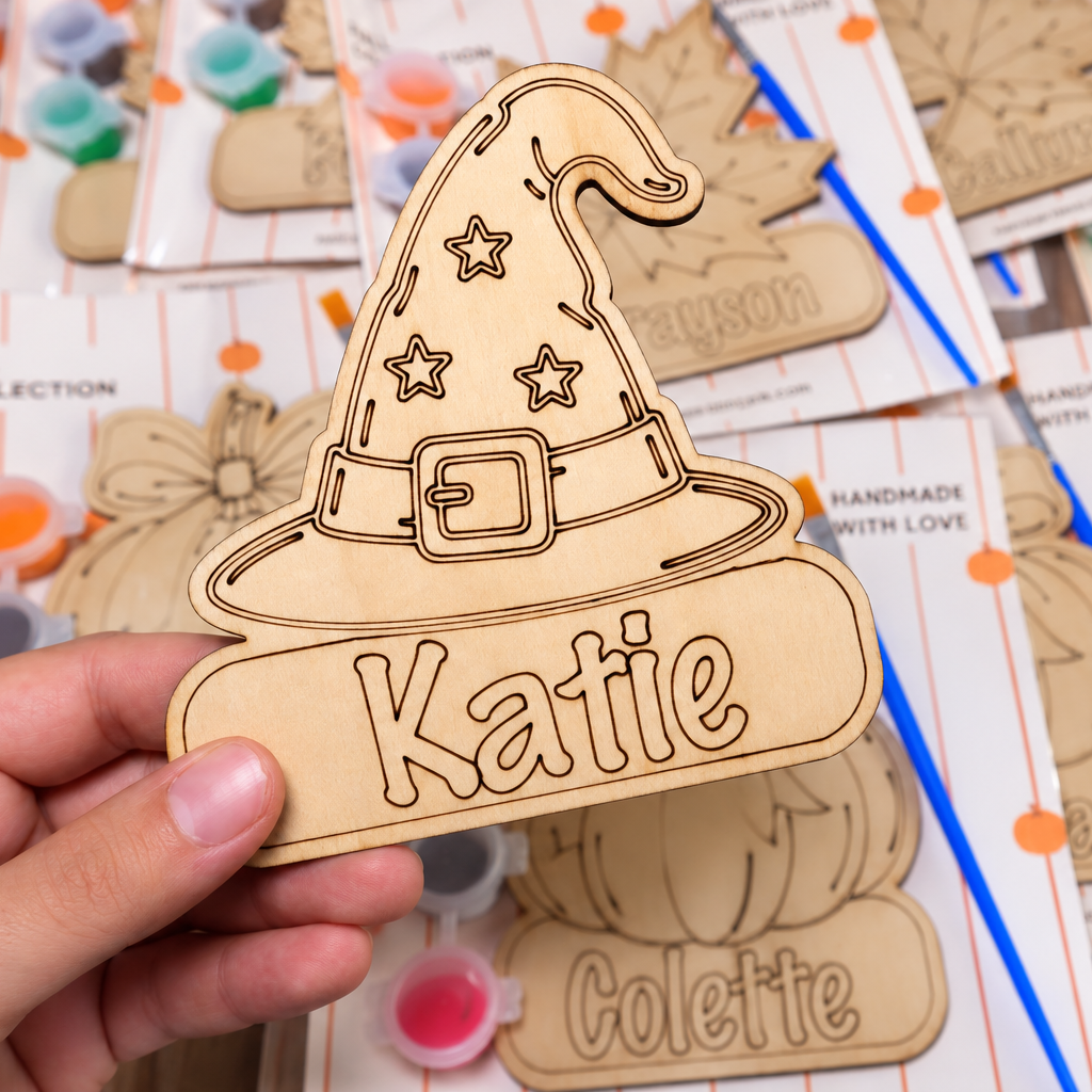 A hand holds a personalized wooden wizard hat cutout engraved with the name 'Katie' on a table covered with similar unfinished wooden craft cutouts and paint pots.