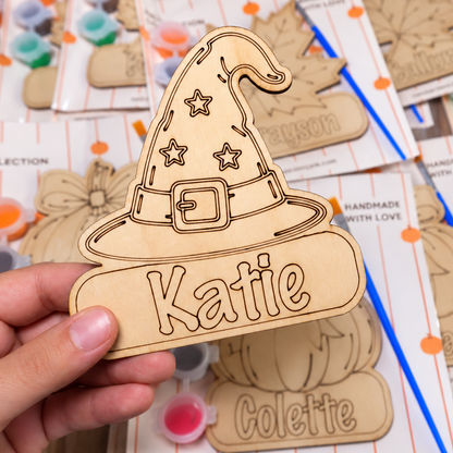A hand holds a personalized wooden wizard hat cutout engraved with the name 'Katie' on a table covered with similar unfinished wooden craft cutouts and paint pots.