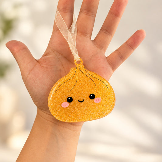 Gold glitter dumpling acrylic ornament with a cute face and sheer ribbon held in a hand.
