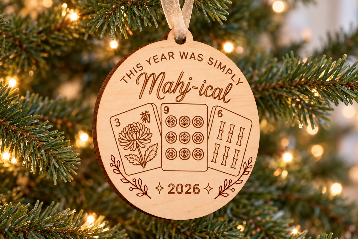 Mahj-ical 2026 engraved wooden mahjong ornament hanging by a beige ribbon on a Christmas tree with warm lights.