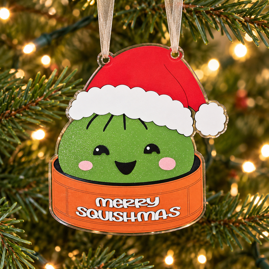 Green merry squishmas UV printed acrylic Christmas ornament hanging on a Christmas tree, with sheer beige ribbon.